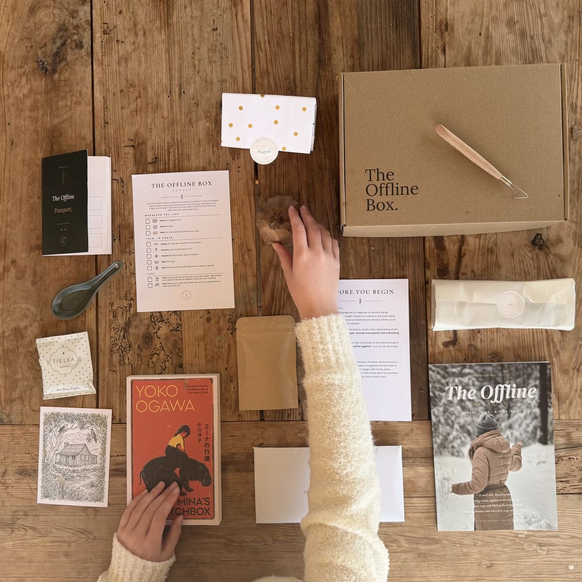 An Offline Box laid out on an oak table: the box, a novel, The Offline magazine, a paint set and small projects, with a hand reaching in